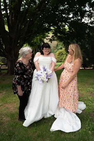 Bride in white off-shoulder gown holding purple bouquet standing with two women under trees outdoors.