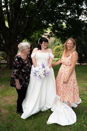Bride in white gown holding purple bouquet poses with two women in a lush green park setting.