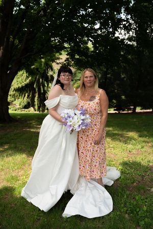 Bride in white off-shoulder gown holding purple bouquet poses with woman in floral dress at outdoor wedding.