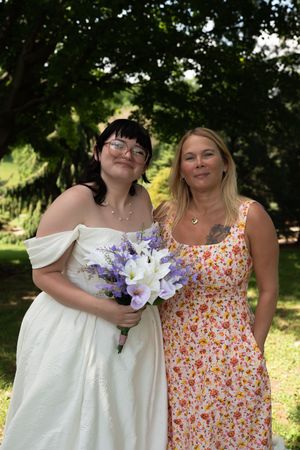 A bride in a white off-shoulder gown holds purple flowers beside a woman in a floral dress outdoors.