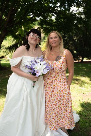 Bride in white off-shoulder gown holding purple and white bouquet poses with smiling woman in floral dress outdoors.