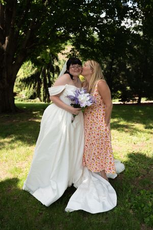 Bride in white off-shoulder gown holding purple bouquet receives kiss on cheek from woman in floral dress outdoors.