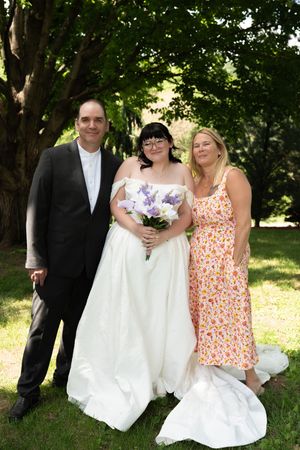 Bride in white gown holding purple bouquet poses with two guests under lush green trees at outdoor wedding.