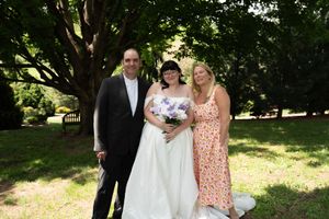 Bride in white gown with purple bouquet poses with man in suit and blonde woman outdoors.