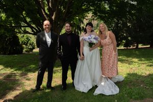 Wedding party of four poses outdoors under green trees with bride in white gown holding purple floral bouquet.