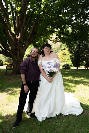 Bride in white off-shoulder gown holds purple bouquet beside groom in black suit outdoors.