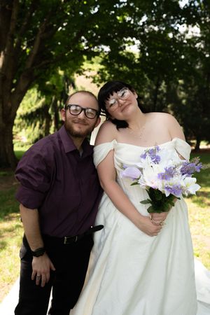 Newlywed couple poses outdoors, bride in white gown holds purple and white bouquet, groom in maroon shirt.