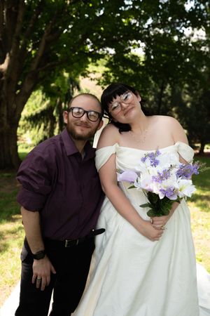 Smiling bride in white off-shoulder gown holds purple bouquet while posing with groom in purple shirt outdoors.