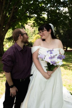 Groom in purple shirt gazes lovingly at bride in white off-shoulder gown holding purple and white bouquet outdoors.