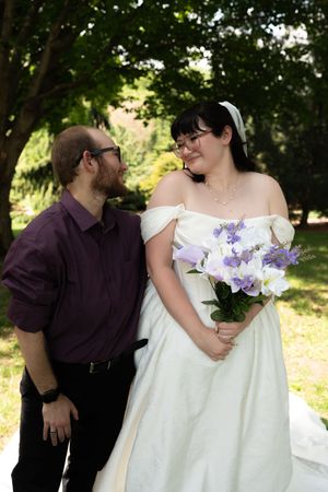 A groom in a purple shirt gazes at his bride holding a bouquet of purple and white flowers outdoors.