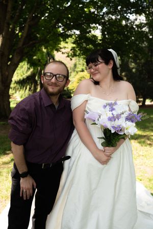 Smiling bride in white gown holds purple bouquet beside groom in purple shirt outdoors.