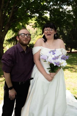 Bride in white off-shoulder gown holds purple bouquet posing with guest in maroon shirt.