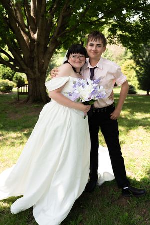 Bride in white off-shoulder wedding gown embraces young man in pink shirt and tie at outdoor wedding celebration.