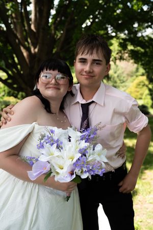 Bride in white dress holding purple and white bouquet posing with young man in pink shirt.