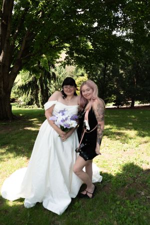 Bride in white off-shoulder gown holds purple bouquet posing with guest in black dress outdoors.