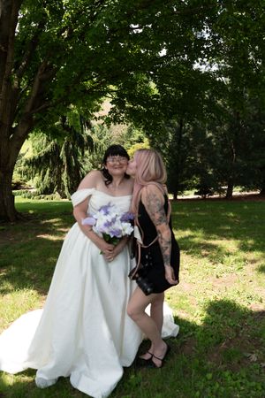 Tattooed woman in black dress kisses bride in white off-shoulder gown in garden setting.