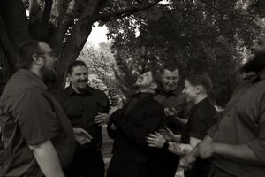 Black and white photo of groom laughing joyfully surrounded by groomsmen in black attire under shaded trees.