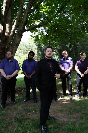 Groomsmen in purple shirts stand behind groom in black suit outdoors under green trees.