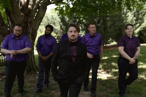 Five groomsmen in matching purple shirts and black pants pose together under a tree at an outdoor wedding.
