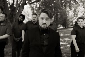 Black and white group portrait of a groom and groomsmen all dressed in black standing among trees.