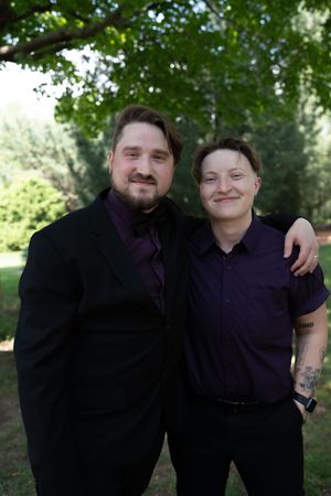 Two men in matching purple shirts and black suits smile together in a green park setting.