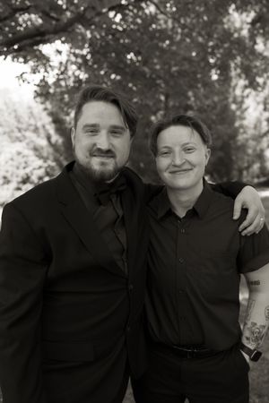 Black and white photo of two people in formal black attire smiling together outdoors at a wedding.