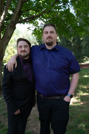 Two men in formal attire pose together outdoors under green trees at a wedding celebration.