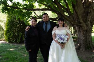 Bride in white gown holding purple bouquet poses with groom and officiant under large tree at outdoor wedding.