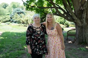 Two women stand together smiling in a lush green garden park setting on a sunny day.
