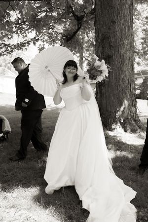 Black and white photo of bride in white gown holding parasol and bouquet under trees.