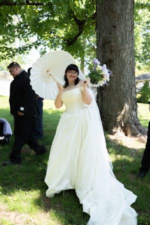 Bride in white gown holds parasol and bouquet under green trees in outdoor setting.