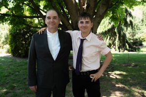 Two men in formal attire smiling together outdoors under a large tree.