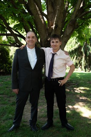 Two males in formal suits posing together outdoors near a large tree at a wedding celebration.