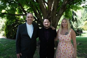 Three adults posing together outdoors under a large tree at a wedding celebration.