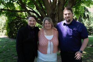 Two men in purple shirts flank a smiling woman in pink at an outdoor wedding celebration.