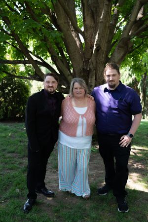 Two men in formal attire pose with smiling woman under large tree in garden.