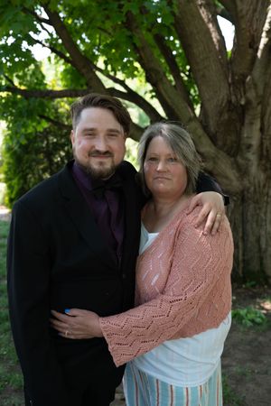 Man in black suit with purple tie embraces woman in pink cardigan under large tree at wedding.