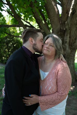 Man in black suit gently kisses woman in pink cardigan on cheek under green tree outdoors.