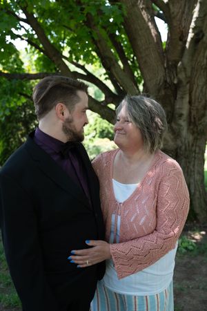 Couple in wedding attire gazes lovingly at each other beneath large tree in outdoor garden setting.