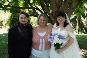 Bride in white gown with purple bouquet poses with groom and woman under large tree.