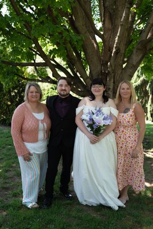 Wedding party poses under large tree, bride in white gown holding purple bouquet with groom and two guests.