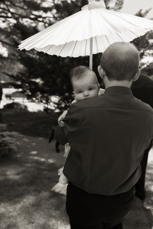Man holds baby under white parasol umbrella in black and white outdoor wedding photo.