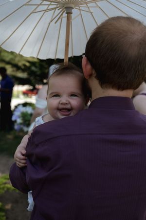 Smiling baby being held by a man in a purple shirt at an outdoor wedding ceremony under a white parasol.