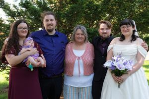 Family group of five poses with bride in white gown and purple bouquet at outdoor wedding celebration.