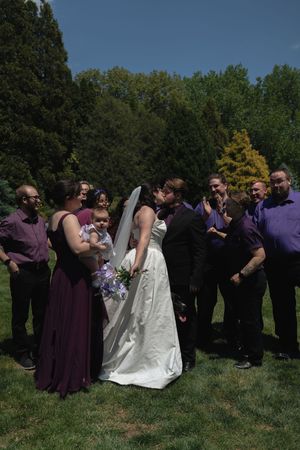 Wedding party gathered outdoors as bride and groom share first kiss surrounded by guests in purple attire.