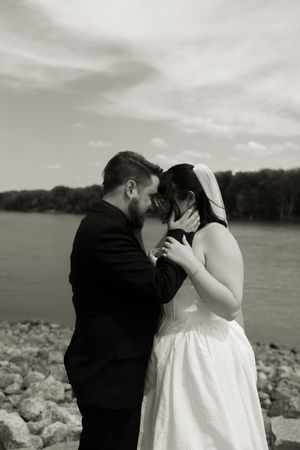 Black and white romantic photo of bride and groom touching foreheads near a river.