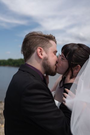 Bride and groom share romantic first kiss by lakeside on sunny wedding day, veil flowing in the breeze.