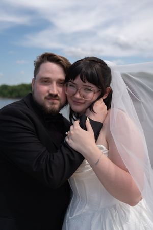 A bride and groom embrace tenderly outdoors near water, she wearing a white veil and glasses, he in a black suit.