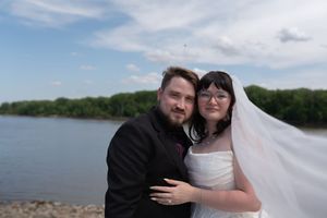 Bride and groom embrace on rocky rivershore, her veil flowing in the breeze under blue sky.