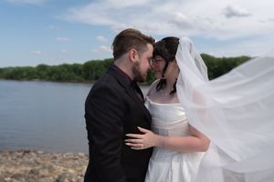 Bride and groom share a romantic moment by the river, her veil billowing in the wind.